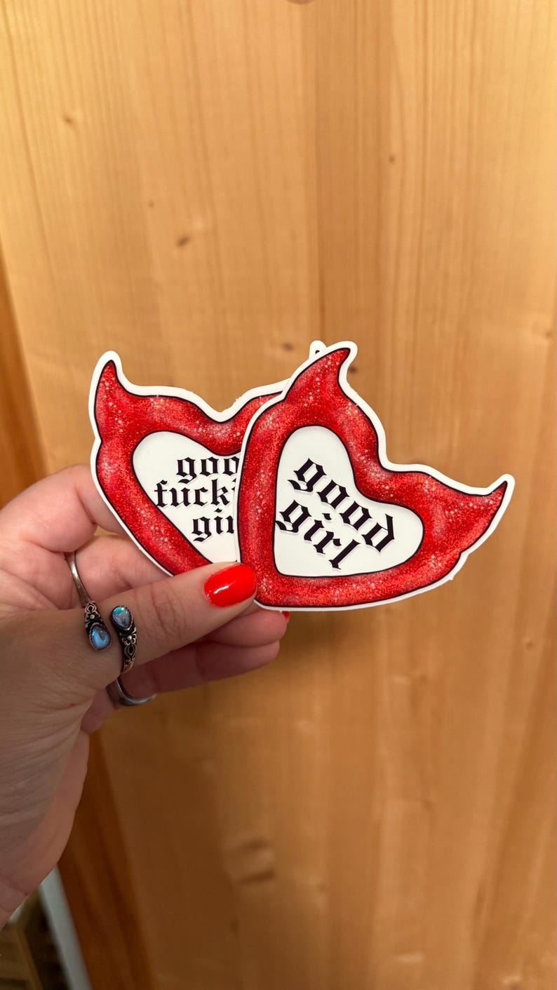 May include: Two heart-shaped stickers with red glitter borders. One sticker reads "good girl" in gothic font, the other reads "god fuckin' girl". The stickers are held in a hand with red nail polish and a silver ring.