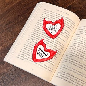 May include: Two red heart-shaped stickers with the words "good fucking girl" and "good girl" in a gothic font are placed on an open book. The stickers have a white border and a devil-like shape.