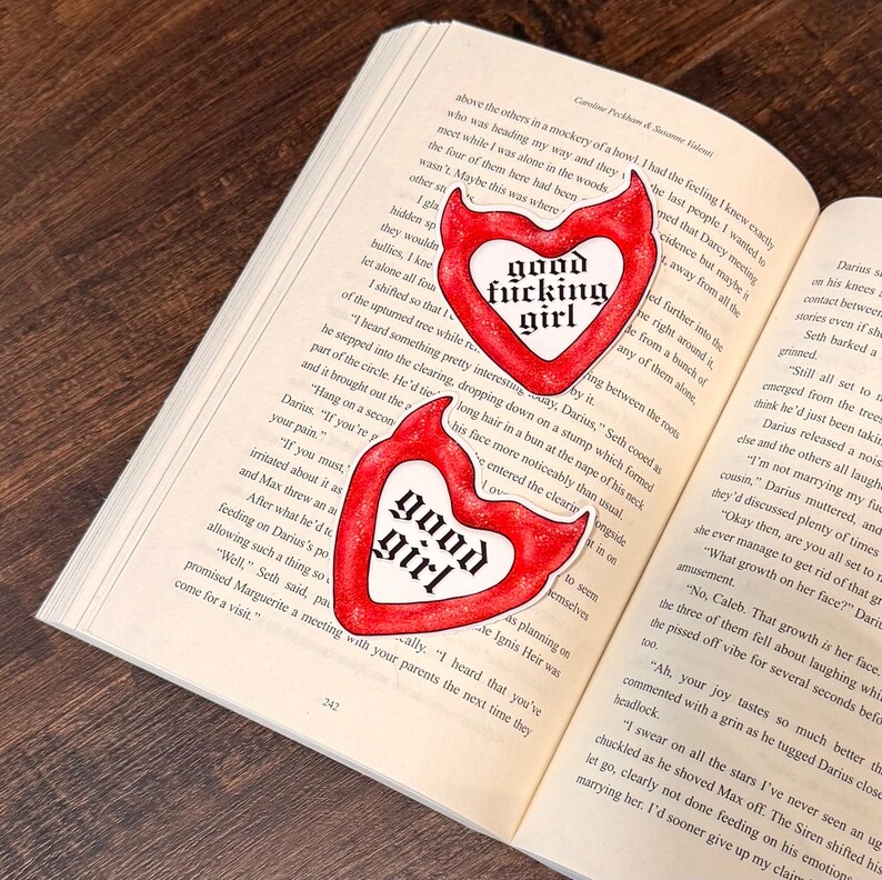 May include: Two red heart-shaped stickers with the words "good fucking girl" and "good girl" in a gothic font are placed on an open book. The stickers have a white border and a devil-like shape.