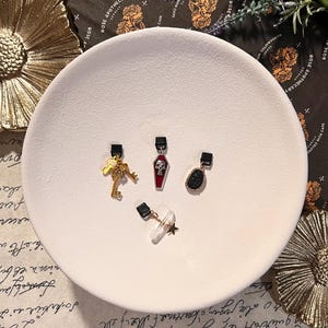 May include: A selection of decorative charms displayed on a white plate. The charms feature a gold key, a coffin with a skull, a black stone with a silver border, and a crystal with a gold star. Each charm has a black clip.