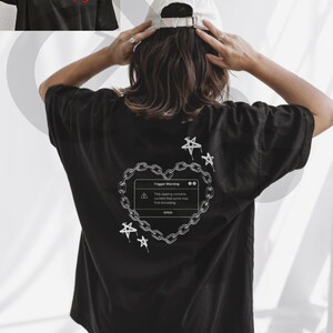 May include: Black t-shirt with a white graphic on the back featuring a heart-shaped chain design with a pop-up message that reads "Trigger Warning". The front of the shirt has a small red rose design.