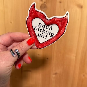 May include: A red heart-shaped sticker with glitter accents and the words "good fucking girl" in a gothic font. The sticker is held against a wooden background.