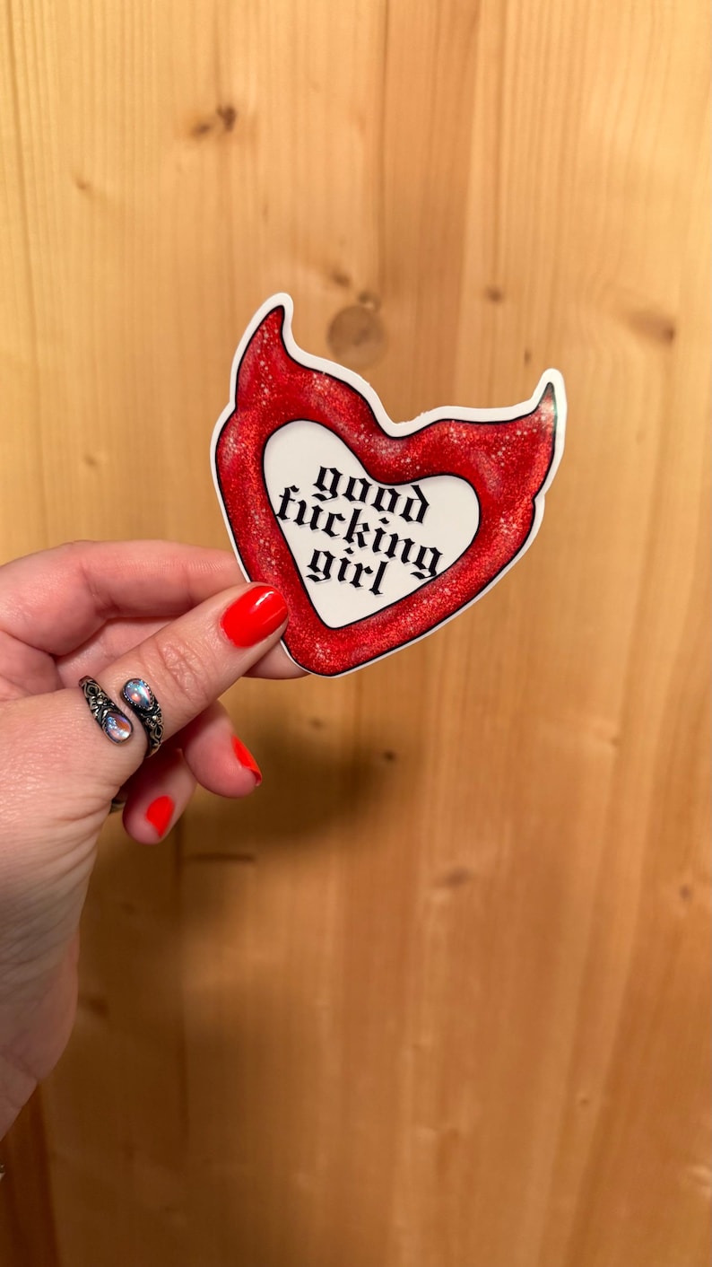 May include: A red heart-shaped sticker with glitter accents and the words "good fucking girl" in a gothic font. The sticker is held against a wooden background.
