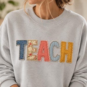 Pode incluir: Uma camisola de gola redonda cinzento-claro com a palavra TEACH aplicada no peito. Cada letra é feita de um tecido diferente: ganga azul, estampado floral, coral sólido, riscas azuis e brancas e amarelo mostarda sólido.