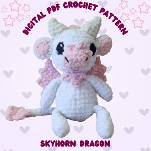 Op de afbeelding: Een wit en roze gehaakte drakenknuffel met zwarte ogen, roze vleugels en een roze snuit. De tekst "DIGITAL PDF CROCHET PATTERN" en "SKYHORN DRAGON" staan in de afbeelding. De draak heeft een sjaal om zijn nek.