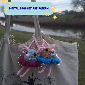 Op de afbeelding: Twee gehaakte dierenbedels, één grijs en één bruin, met tutu's en roze bandjes, hangen aan een beige draagtas. De bedels hebben kleine oren en zijn met roze garen aan de tas bevestigd. De tekst bovenaan luidt "DIGITAL CROCHET PDF PATTERN."