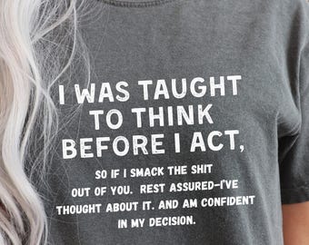 May include: Dark grey t-shirt with white text. The text reads: "I WAS TAUGHT TO THINK BEFORE I ACT, SO IF I SMACK THE SHIT OUT OF YOU. REST ASSURED-I'VE THOUGHT ABOUT IT. AND AM CONFIDENT IN MY DECISION."