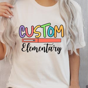 Pode incluir: Camiseta branca com a palavra "CUSTOM" em letras coloridas, uma barra de carregamento e a palavra "Elementary" em fonte cursiva. A camiseta é de estilo casual.