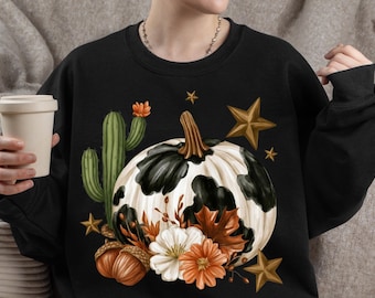 May include: A black long-sleeve sweatshirt featuring a graphic print of a white pumpkin with black cow-print spots, a green cactus, brown acorns, and orange and white flowers. The design is accented with several gold-colored stars.