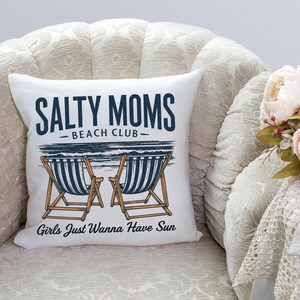May include: White cushion with the text "Salty Moms Beach Club" and "Girls Just Wanna Have Sun" in black and blue lettering. The cushion features a graphic of two deckchairs with blue and white stripes.
