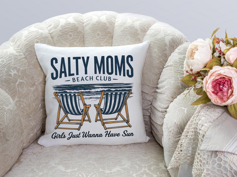 May include: White cushion with the text "Salty Moms Beach Club" and "Girls Just Wanna Have Sun" in black and blue lettering. The cushion features a graphic of two deckchairs with blue and white stripes.