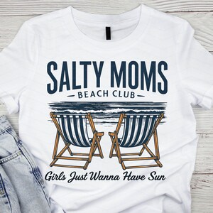 May include: White t-shirt with a blue graphic that says "Salty Moms Beach Club" and features two beach chairs with a blue and white striped design. The text "Girls Just Wanna Have Sun" is below the chairs.