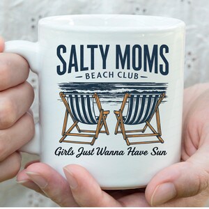 May include: White ceramic mug with a black and white illustration of beach chairs and the text "Salty Moms Beach Club - Girls Just Wanna Have Sun".