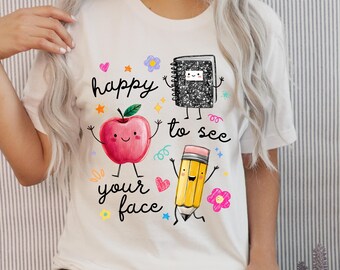 May include: A white t-shirt featuring a graphic print of a red apple, a black and white composition notebook, and a yellow pencil, all with smiling faces and limbs. The text "happy to see your face" is printed in black script among colorful stars and flowers.
