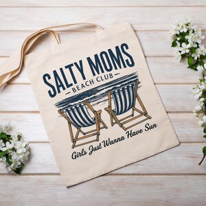 May include: A white canvas tote bag with the text "Salty Moms - Beach Club" and an illustration of two deckchairs. The text below the chairs reads "Girls Just Wanna Have Sun."