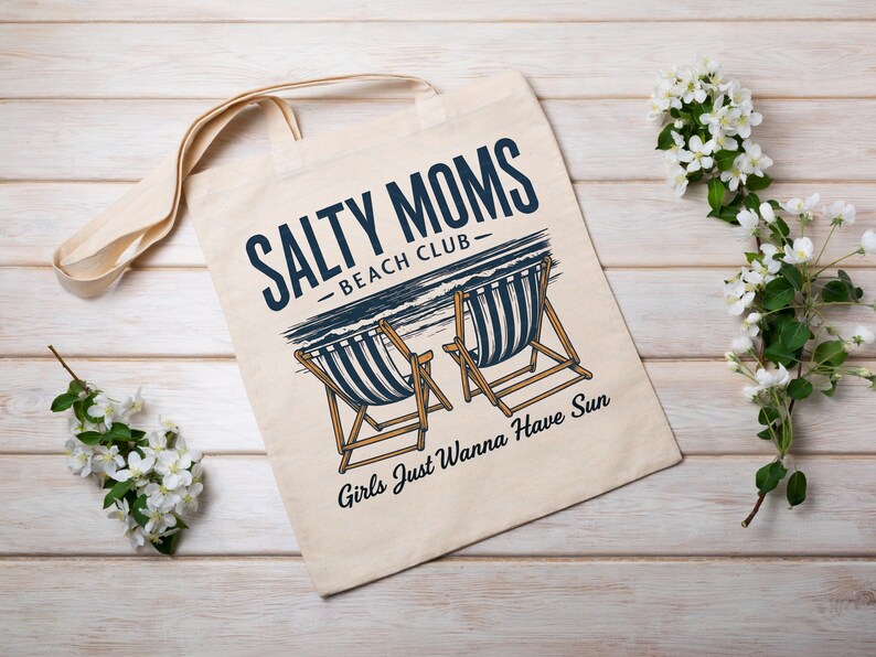 May include: A white canvas tote bag with the text "Salty Moms - Beach Club" and an illustration of two deckchairs. The text below the chairs reads "Girls Just Wanna Have Sun."