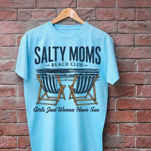 May include: Light blue t-shirt with a graphic design of two beach chairs and the text "Salty Moms Beach Club" and "Girls Just Wanna Have Sun".