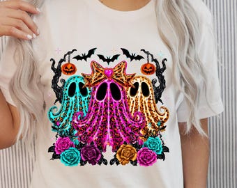May include: A white t-shirt featuring a graphic print of three ghosts with leopard print patterns in turquoise, pink, and orange. The design includes black bats, hanging jack-o'-lanterns, and colorful roses at the base of the ghosts.