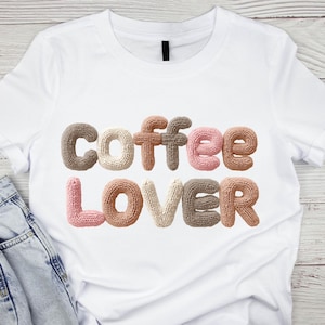 Könnte beinhalten: Weißes T-Shirt mit dem Schriftzug "COFFEE LOVER" in einer gestrickten, strukturierten Schriftart. Die Buchstaben sind in Braun-, Rosa- und Beigetönen gehalten. Das Shirt wird auf einer hellen Holzoberfläche mit einer Jeans präsentiert.
