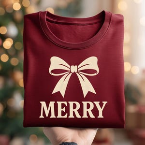 Könnte beinhalten: Ein gefaltetes weinrotes T-Shirt mit einer cremefarbenen Schleife und dem Wort "MERRY" darunter. Das Shirt wird vor einem verschwommenen Hintergrund aus Weihnachtslichtern gehalten.