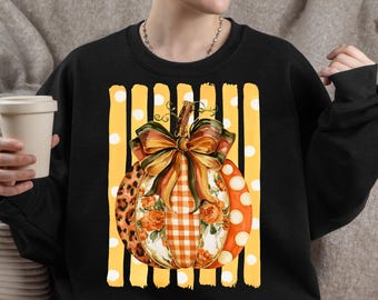 May include: A black long-sleeve sweatshirt featuring a central graphic of a pumpkin decorated with a large bow. The pumpkin has sections with leopard print, orange gingham, and floral patterns. It sits against a yellow background with white stripes.