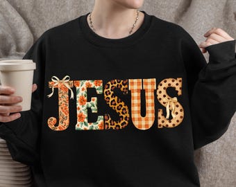 May include: A black long-sleeve sweatshirt featuring the word JESUS in large block letters. Each letter has a unique pattern: floral, leopard print, orange gingham, and polka dots. The J and S are decorated with small, light-colored ribbon bows.
