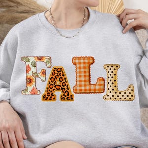 May include: A light gray crewneck sweatshirt featuring the word FALL printed across the chest. Each letter has a distinct pattern: the F features sunflowers and leaves, the A has a leopard print, the L is orange gingham, and the L has black dots.