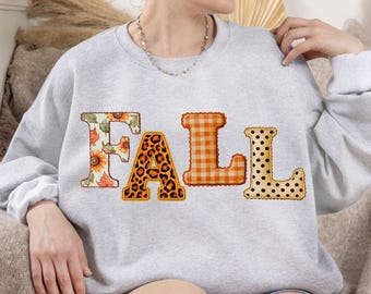 May include: A light gray crewneck sweatshirt featuring the word FALL printed across the chest. Each letter has a distinct pattern: the F features sunflowers and leaves, the A has a leopard print, the L is orange gingham, and the L has black dots.