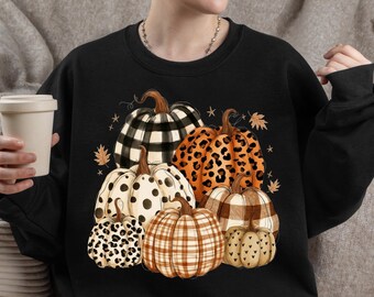 May include: A black long-sleeve sweatshirt featuring a graphic print of six pumpkins with various patterns including black and white buffalo plaid, orange leopard print, white with black polka dots, brown plaid, and small black hearts on a tan pumpkin.