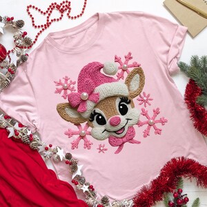 Peut inclure: Un t-shirt rose clair avec un motif de renne portant un bonnet et un nœud roses en tricot, entouré de flocons de neige roses. Le renne a de grands yeux et un nez rose. Le t-shirt est orné d'un accent de perle.