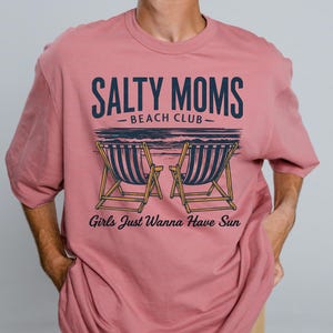May include: A pink t-shirt with the text "Salty Moms - Beach Club - Girls Just Wanna Have Sun" and a graphic of two deckchairs.