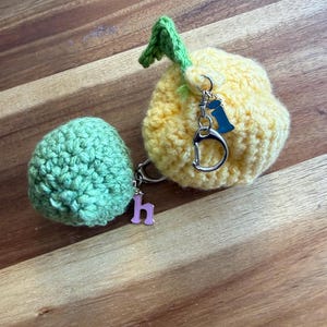 May include: Two crocheted fruit-shaped keyrings. One is yellow with a green stem, the other is green. Each keyring has a silver ring and a charm with a letter. The keyrings are on a wooden surface.