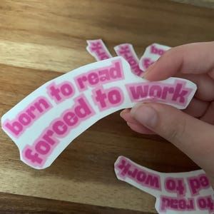 以下が含まれることがあります： 「born to read forced to work」と書かれたピンク色の文字が入った白いステッカー。いくつかのステッカーが見え、一部は手で持たれています。ステッカーは木製の表面にあります。