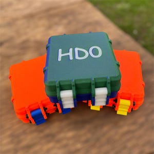Peut inclure: Trois boîtes de rangement en plastique empilables. La boîte du haut est verte avec le texte "HDO" en blanc. La boîte du milieu est bleue et la boîte du bas est orange.