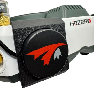 May include: Close-up of a black circular button with a red and white logo on a black and gray headset. The logo is a stylized bird with wings spread. The headset has the text "HDZERO" printed on it.