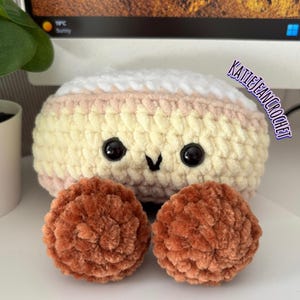 May include: A round, plush crochet toy featuring a white top, a cream-colored middle section, and two textured brown circular feet. The toy has two black plastic eyes and a small black stitched mouth. The text KATIEJEANCROCHET is visible on the right.