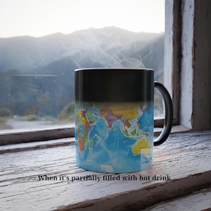May include: A black magic mug with a world map design that appears when filled with a hot beverage. The mug is sitting on a wooden surface in front of a window with a view of mountains.