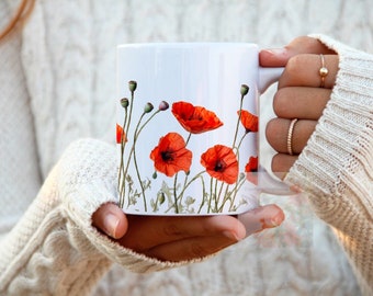 May include: White ceramic mug with a floral design featuring red poppies. The mug is being held by a person wearing a white sweater.