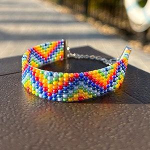 May include: A colorful beaded bracelet with a chevron pattern in shades of blue, green, yellow, orange, and red. The bracelet is made with small, round beads and has a silver clasp.