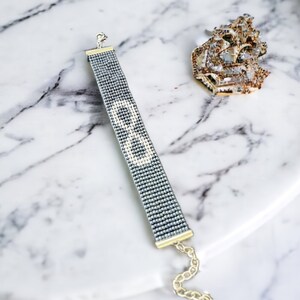 May include: A silver beaded bracelet with a white beaded letter "B" on a marble surface.