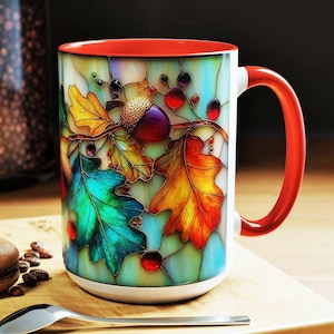 Puede incluir: Una taza de cerámica blanca con interior y asa rojos, con un diseño de vidriera de hojas de otoño y una bellota. Las hojas son en tonos verdes, amarillos y naranjas. Una cuchara y un macaron están sobre una superficie de madera.