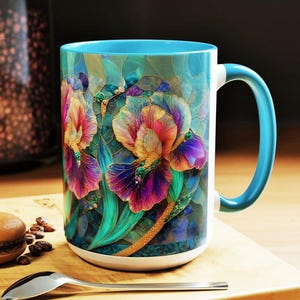 May include: A large ceramic mug with a vibrant floral design featuring irises in shades of purple, orange, and yellow. The mug has a turquoise interior and handle, and is set on a wooden surface with a spoon and a macaron.