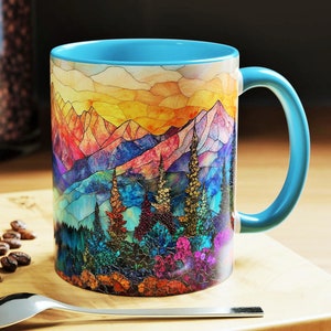 May include: A ceramic coffee mug with a vibrant mountain landscape design. The mug features a light blue interior and handle, and the exterior showcases a colorful mosaic-style illustration of mountains, trees, and a sunset. The mug is sitting on a wooden surface.