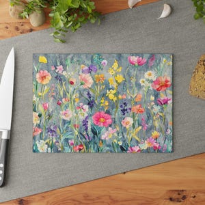 Puede incluir: Tabla de cortar rectangular con un diseño floral en acuarela. La obra de arte presenta una variedad de flores silvestres coloridas en tonos rosa, amarillo, morado y naranja sobre un fondo gris azulado apagado. Un cuchillo de acero inoxidable es visible a la izquierda.