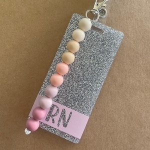 May include: A silver glitter badge holder with a pink and beige beaded retractable badge reel. The badge holder has a pink stripe with the text "RN" on it.