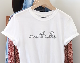 May include: White t-shirt with a black line drawing of a swimmer, cyclist, and runner, with a heartbeat line. The shirt is hanging on a wooden hanger. Other clothing items are visible in the background.