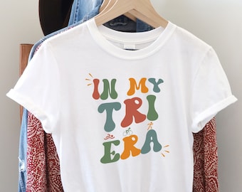 May include: White t-shirt with the text "In My Tri Era" in colorful retro-style lettering. The text is surrounded by small icons representing swimming, cycling, and running.