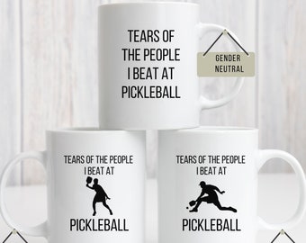 May include: Three white coffee mugs with black text and images. The top mug says "Tears of the people I beat at pickleball" and has a gender neutral label. The middle mug says "Tears of the people I beat at pickleball" and has a female design label. The bottom mug says "Tears of the people I beat at pickleball" and has a male design label.