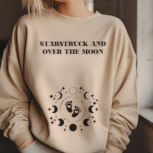 May include: A beige sweatshirt with the text "Starstruck and Over the Moon" printed on the front. Below the text is a graphic of a crescent moon with stars and a pair of baby footprints inside.
