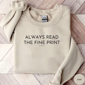 Peut inclure: Un sweat-shirt couleur sable avec le texte "ALWAYS READ THE FINE PRINT" en noir, avec "I'M PREGNANT" en dessous. L'étiquette de la marque Gildan est visible. Le sweat-shirt est plié sur une surface claire imitation bois.